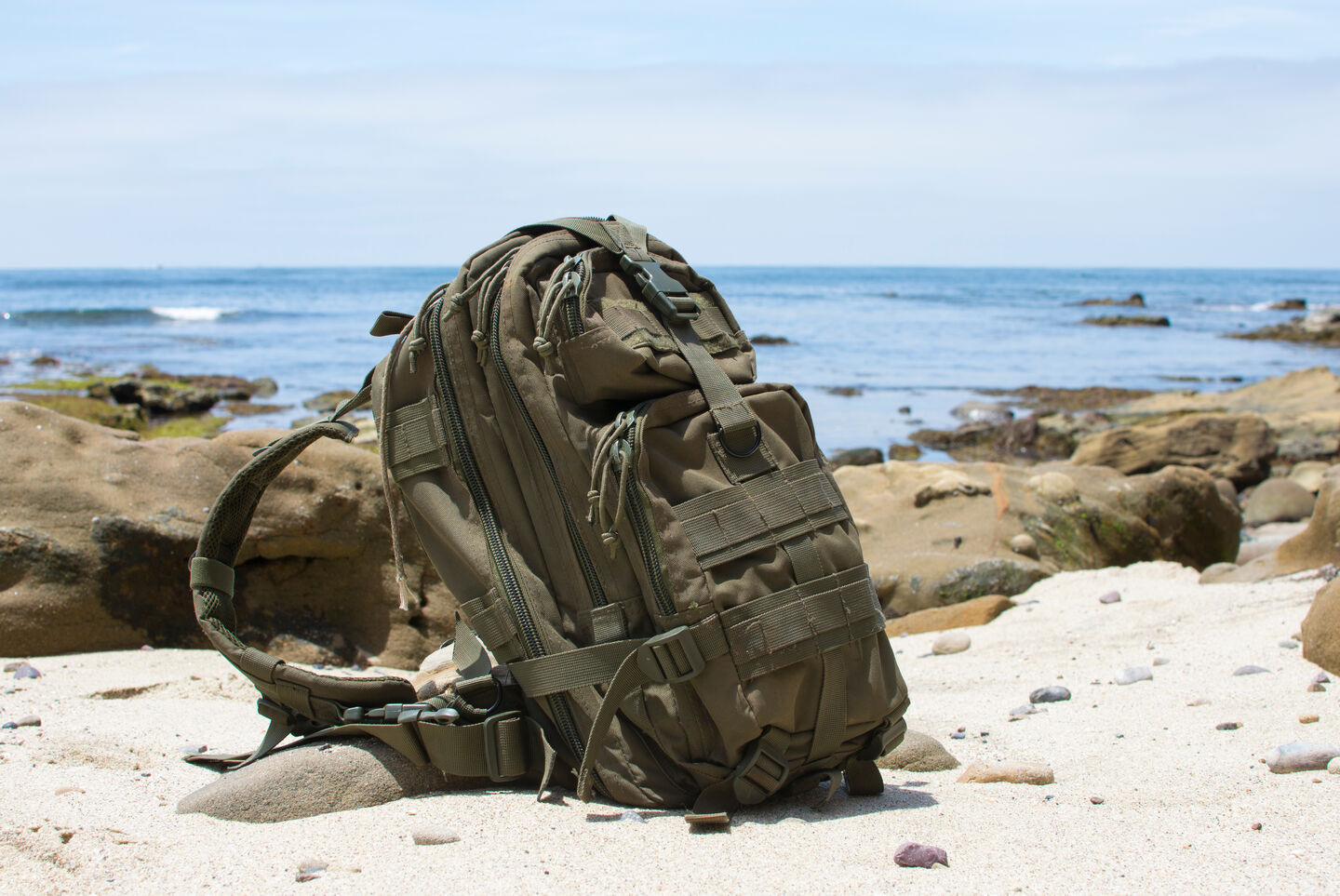 A military green adventure backpack sits upright on the sandy shore of a rocky beach with blue water in the distance.
